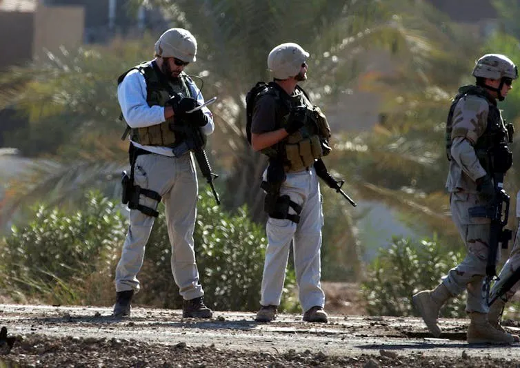 Dos contratistas de seguridad privada estadounidenses investigan el lugar donde un autobús blindado militar fue dañado por una bomba colocada en la carretera cerca del aeropuerto internacional de Bagdad, Irak, en 2004. Créditos: AP